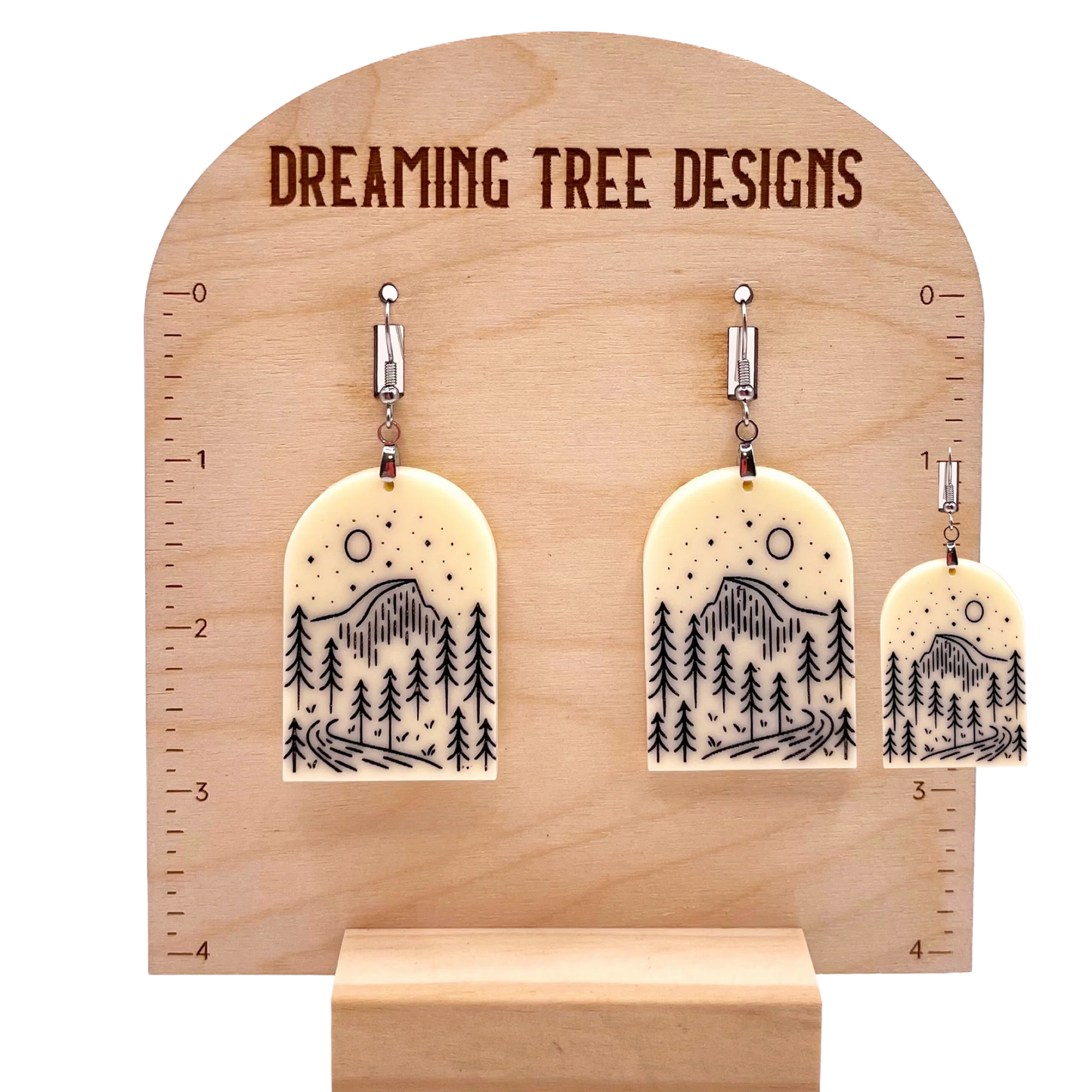 A wooden display stand with a pair of earrings hanging from it. The earrings have a design featuring trees and mountains.