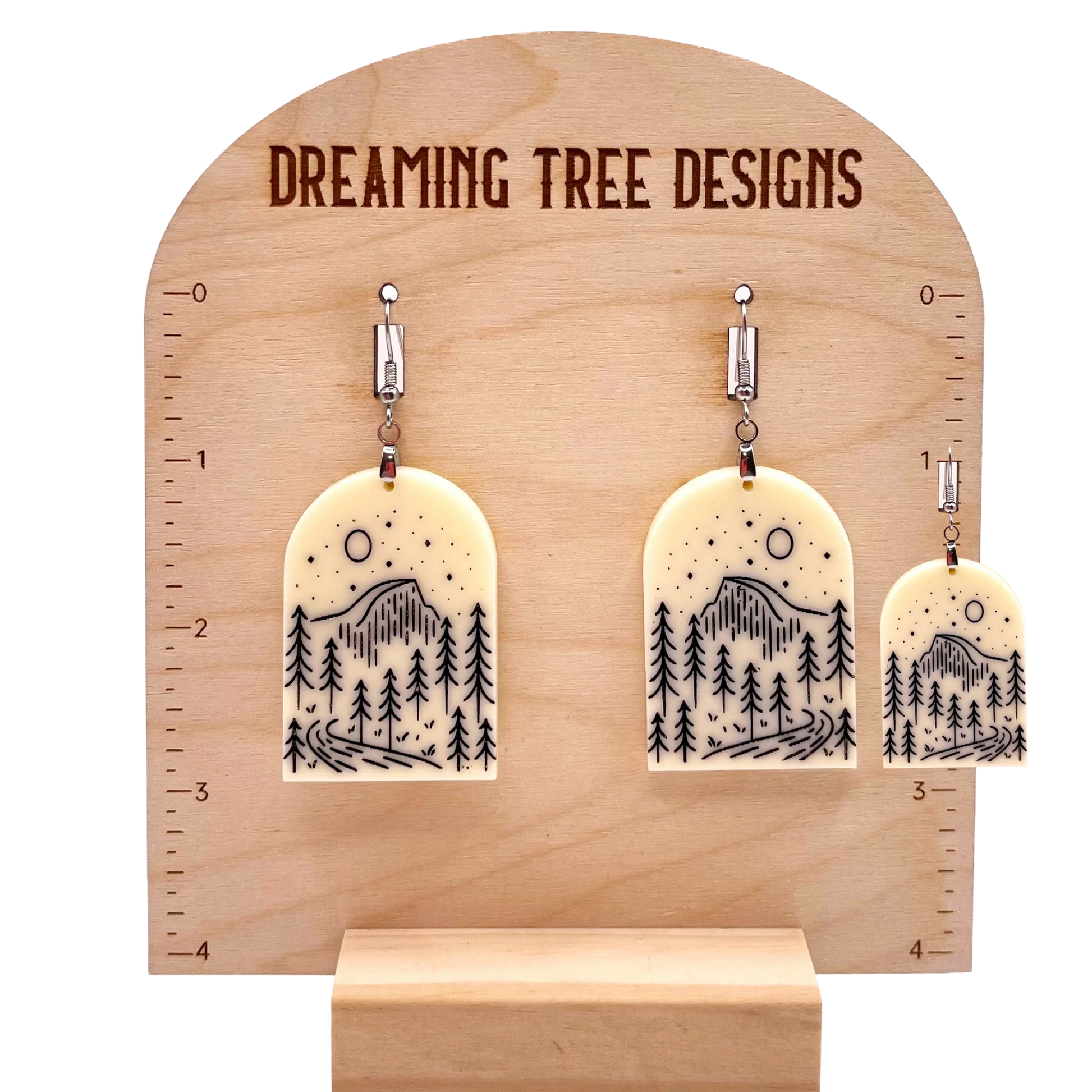 A wooden display stand with a pair of earrings hanging from it. The earrings have a design featuring trees and mountains.